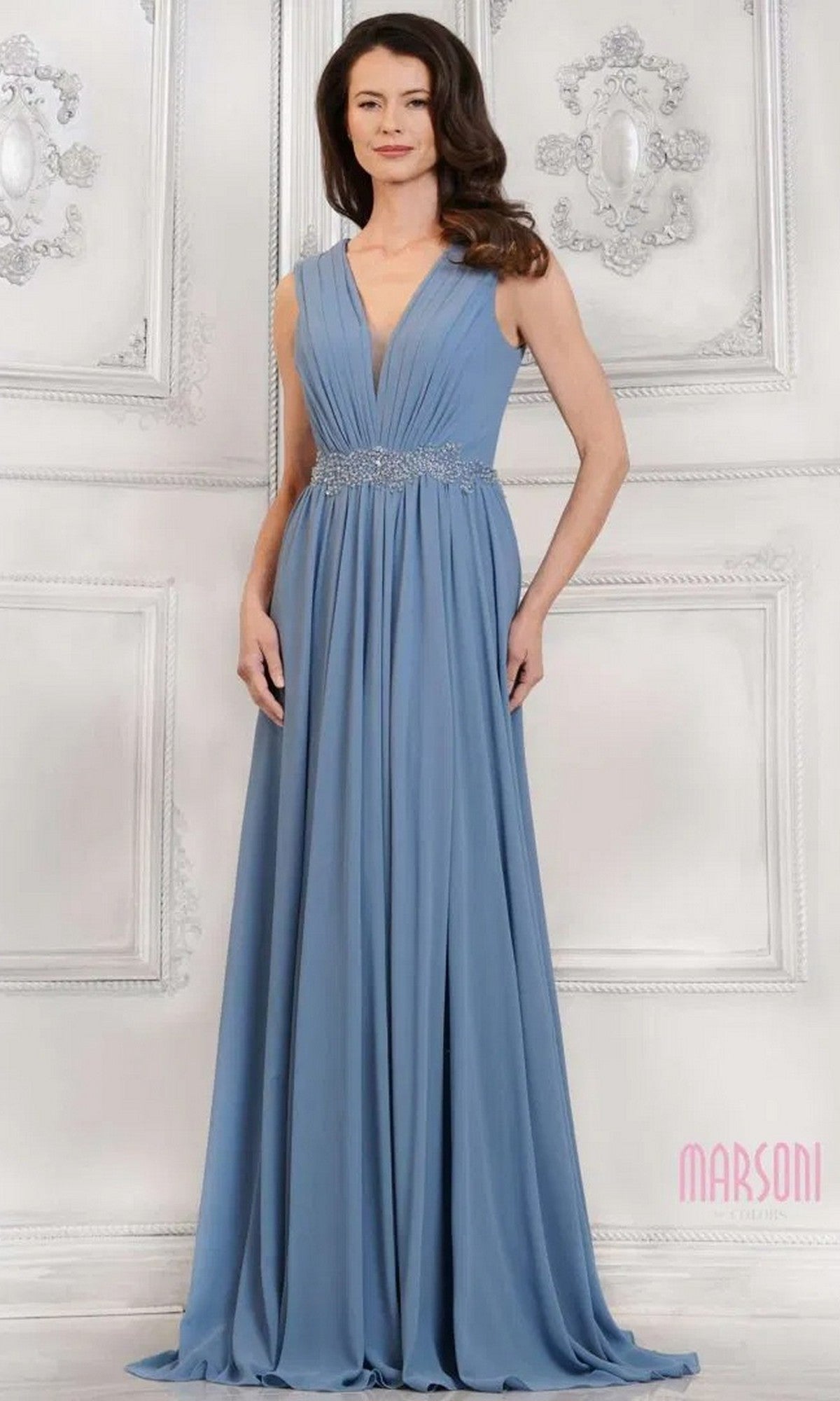 A woman in the Marsoni MV1292 long formal dress in slate blue, featuring a sleeveless design, deep V-neckline, ruched bodice, and beaded waistband, stands before an ornate white wall.